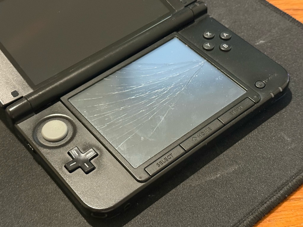 Black 3DS XL with a shattered bottom touch screen, cracks spreading across the digitizer.