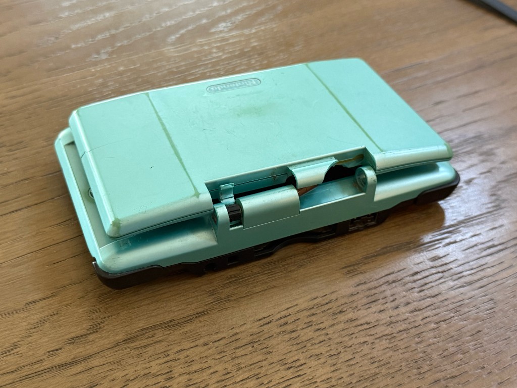 Closed turquoise original DS with the hinge shattered and the top shell split open.