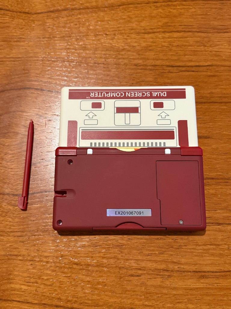 The back of a burgundy and white DS Lite shell printed like a dual screen computer, with a stylus beside it.