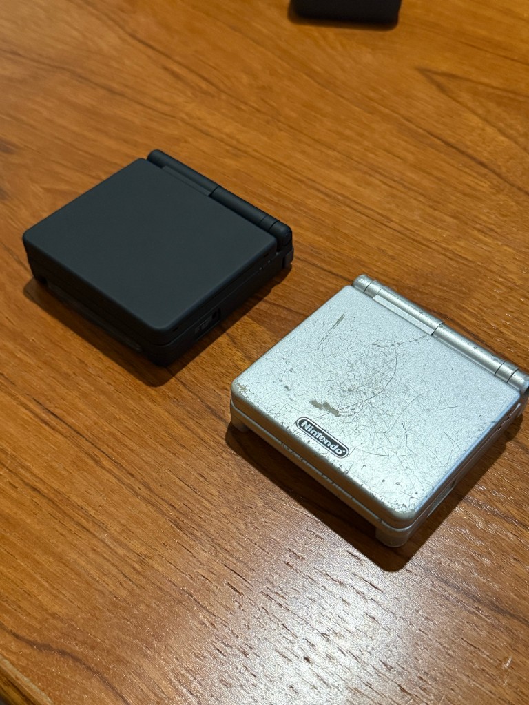 A beat-up silver GBA SP shell next to the matte black shell that replaced it.