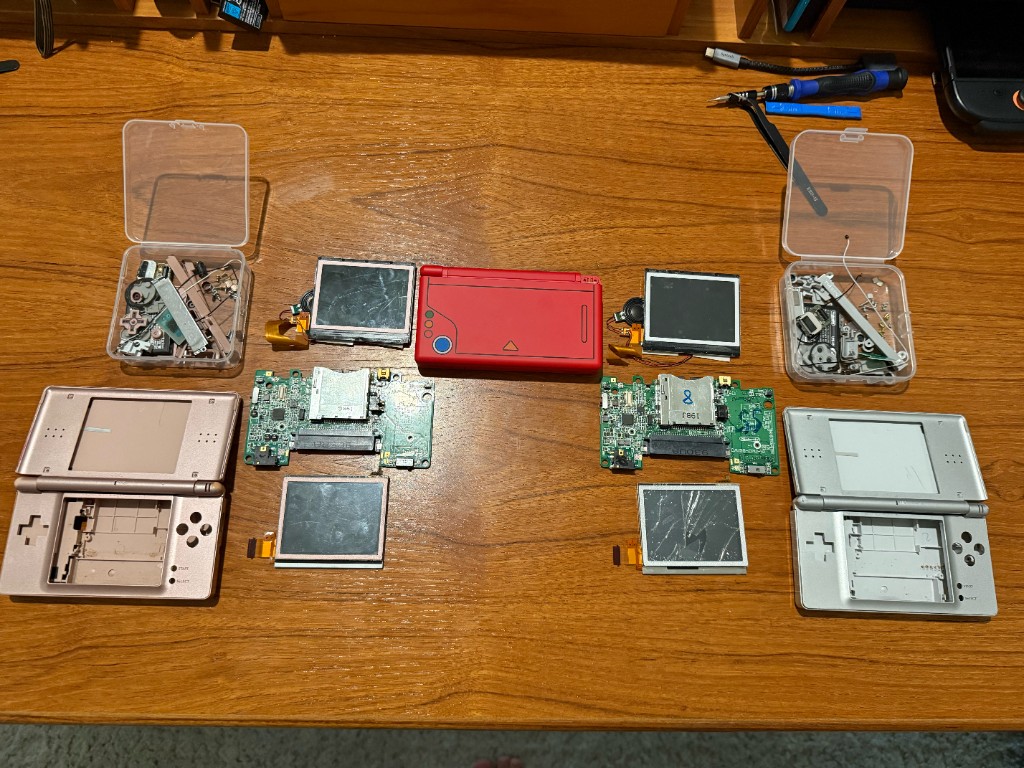 Two DS Lites taken apart on a bench, with cracked screens, boards, shells, and bins of parts.