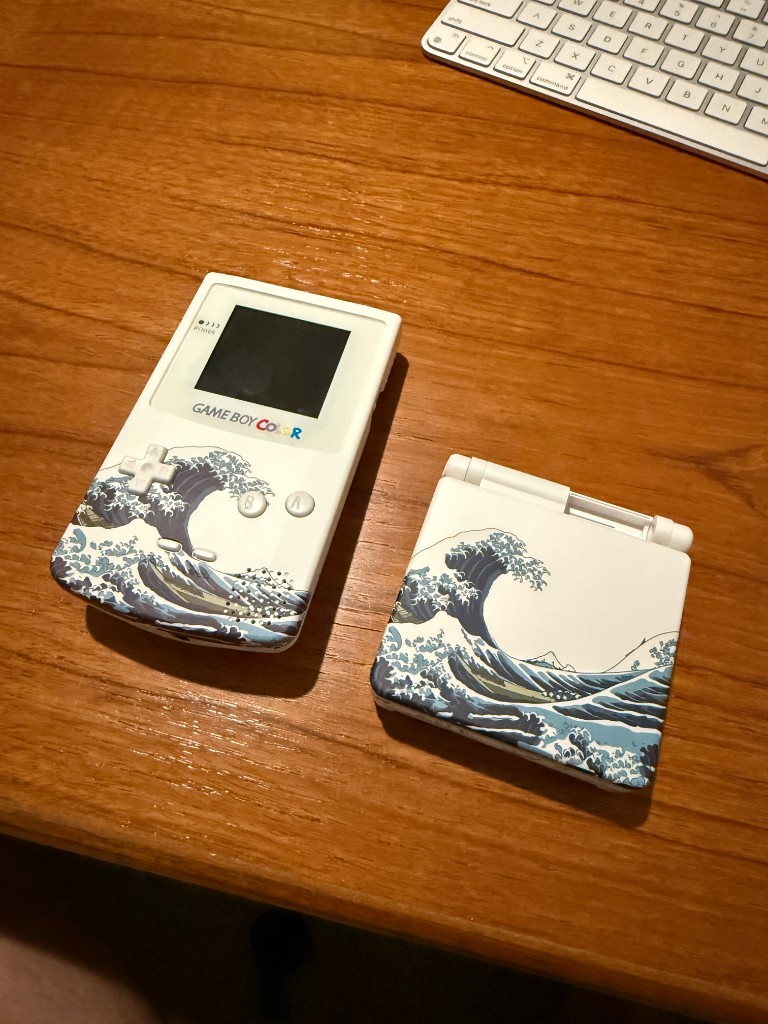 A white Game Boy Color and a white GBA SP, both in shells printed with a matching wave.